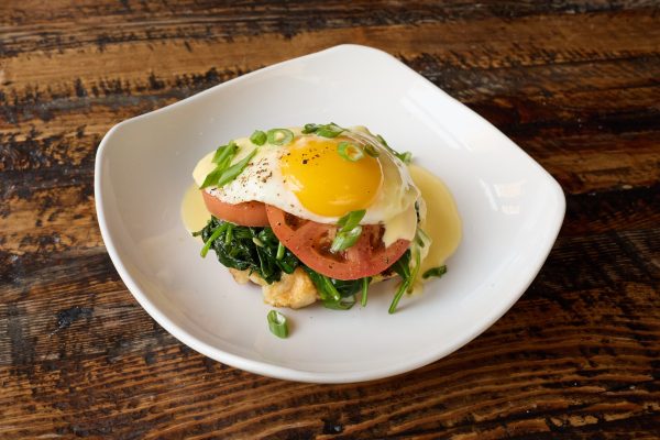 A savory Belgian brunch dish topped with sautéed greens, tomato slices, a sunny-side-up egg, and creamy hollandaise.
