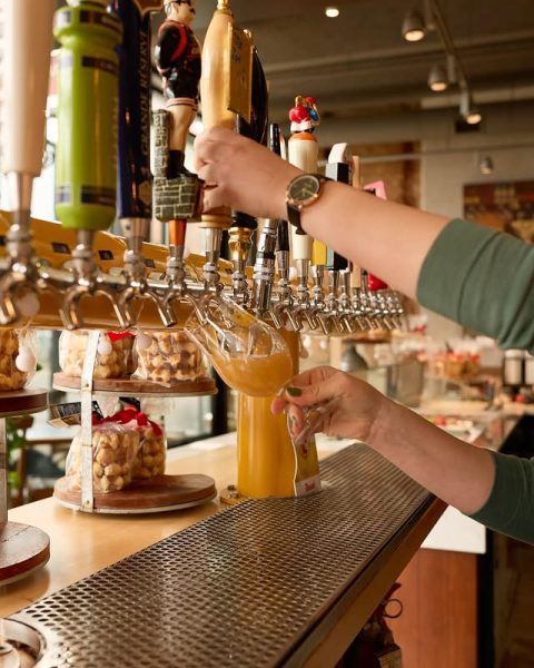 Bartender pouring a draft beer from a colorful tap wall at Taste of Belgium, surrounded by waffle snack displays.