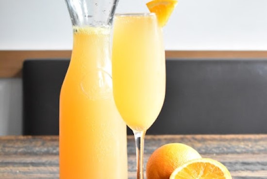 A glass and pitcher of freshly made mimosa garnished with orange slice, beside halved oranges at Taste of Belgium.