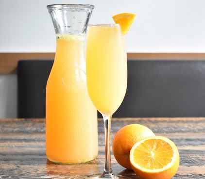 A glass and pitcher of freshly made mimosa garnished with orange slice, beside halved oranges at Taste of Belgium.
