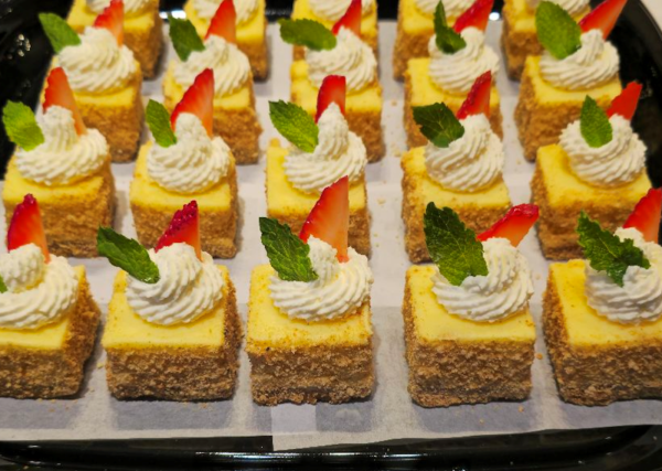 Mini cheesecake bites with a graham cracker crust, topped with whipped cream, strawberry slices, and mint leaves on a tray.