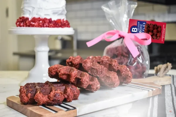 Close-up of Red Velvet Liège waffles displayed on a marble counter next to a frosted cake and packaged waffle gift bag.
